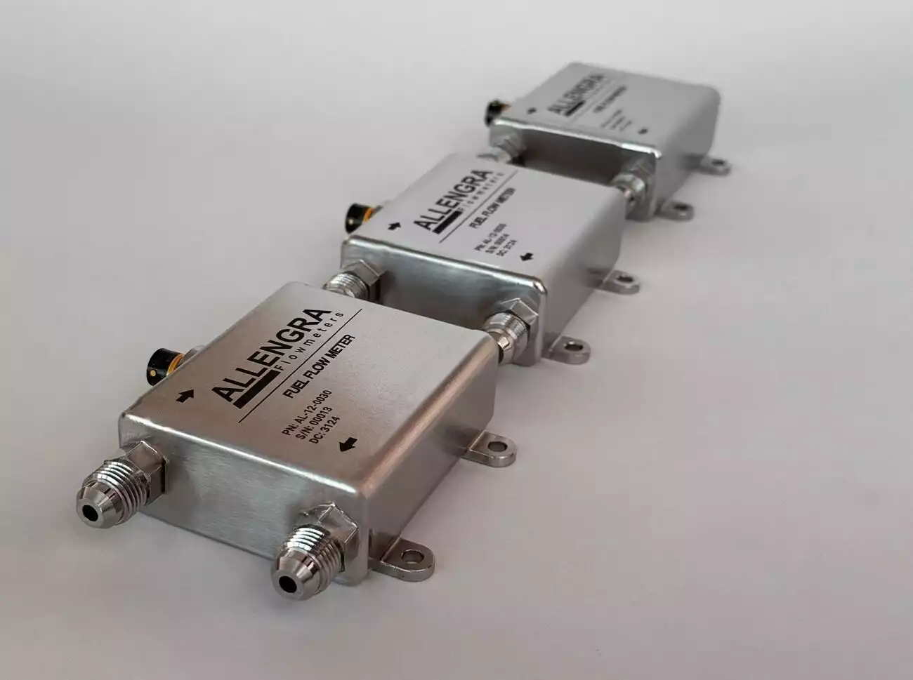 Three Allengra ultrasonic fuel flow meters designed for the 2026 FIA Formula 1 regulations, featuring a silver housing and dual 6AN male connections. These sensors utilize advanced ultrasonic technology to monitor the new 3000 MJ/h energy flow limit and are engineered to withstand the aggressive chemistry of 100% sustainable e-fuels.